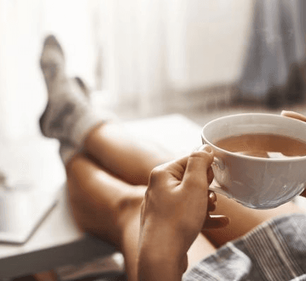 Mãos segurando uma xícara de chá quente enquanto a pessoa relaxa com as pernas cruzadas e apoiadas em uma superfície, com um tablet ou livro por perto. Cenário aconchegante e doméstico.