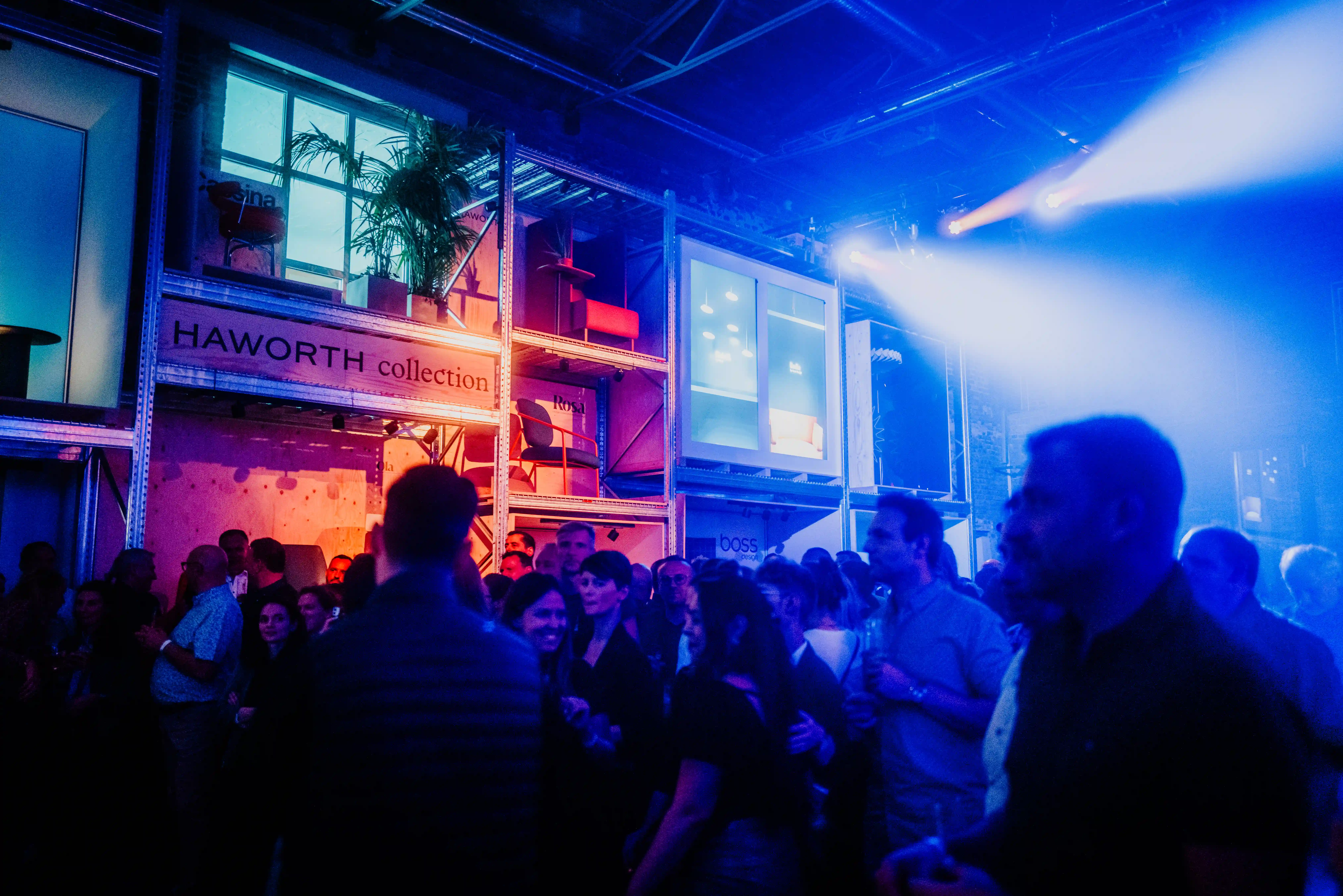 Evening party at the HAWORTH ORGATEC stand with a crowd in blue light, spotlights and the illuminated two-storey Haworth Collection shelf structure.