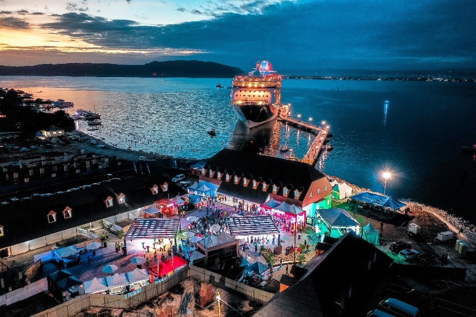 Evening view of a cruise ship docked with a SeaWalk floating pier, at Port Royal in Jamaica, with illuminated waterfront buildings and visitor activity at a coastal destination.