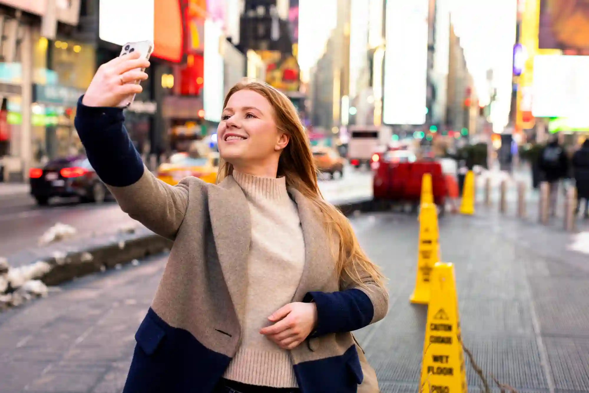 Young woman taking a selfie in a busy city square, likely Times Square, highlighting tourist activities.