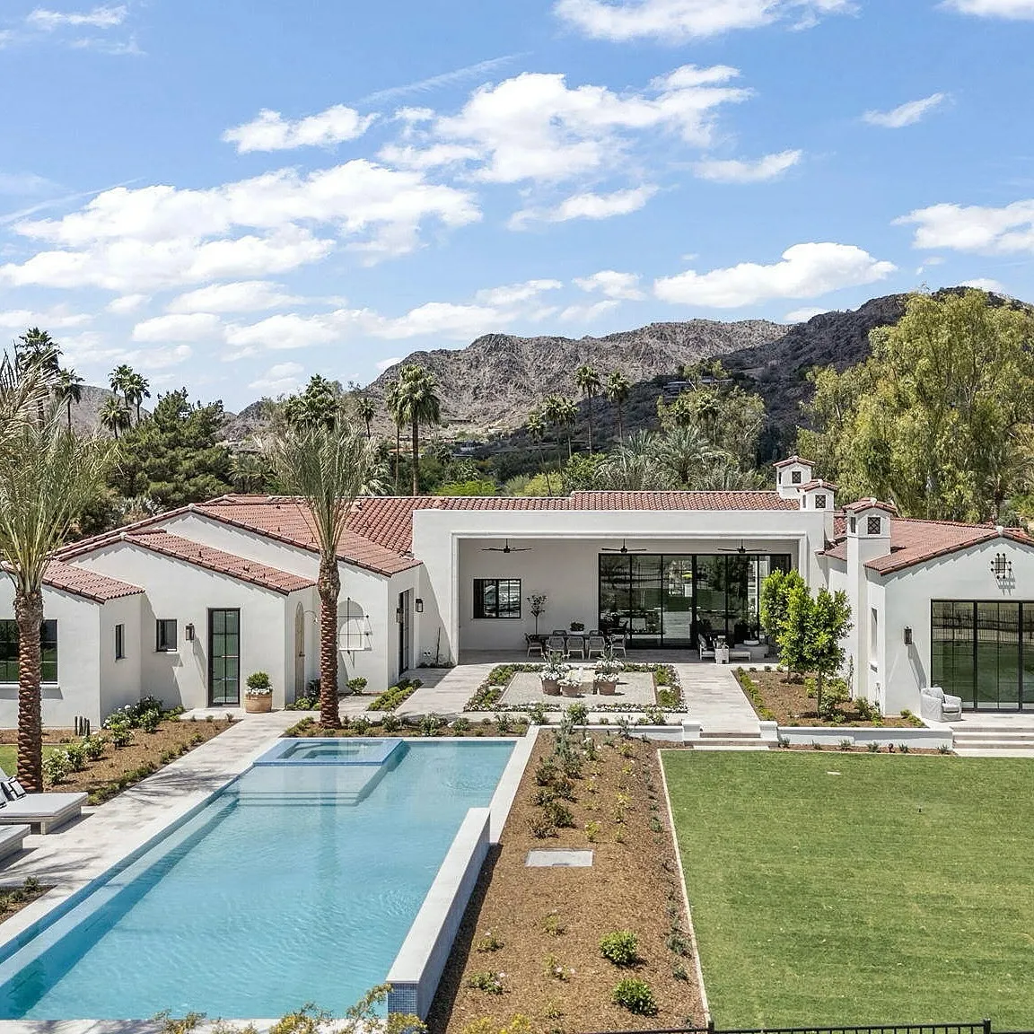 Luxurious home exterior with a tiled roof, infinity pool, green lawn, and mountainous backdrop.