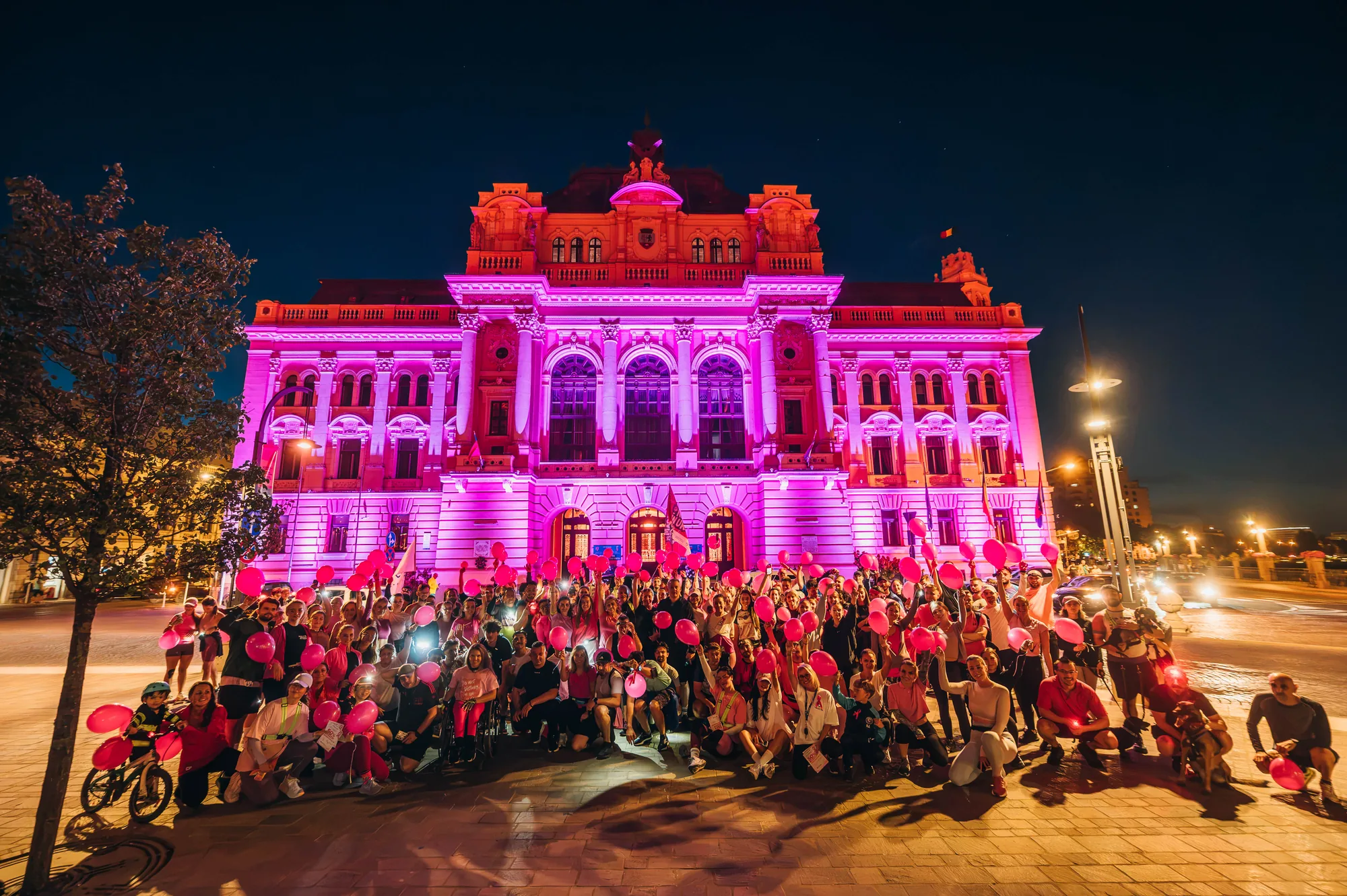 Comunitatea ORC cu baloane roz în fața clădirii iluminate în roz din centrul Oradei.