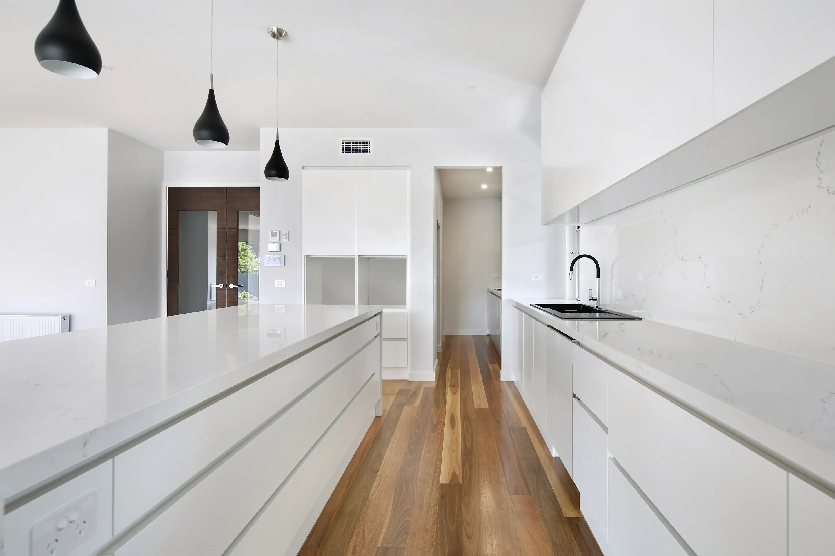 Galley kitchen with white cabinetry, white stone benchtops and splashback, three black pendant lights, a black tap, timber floors and dark timber entry doors