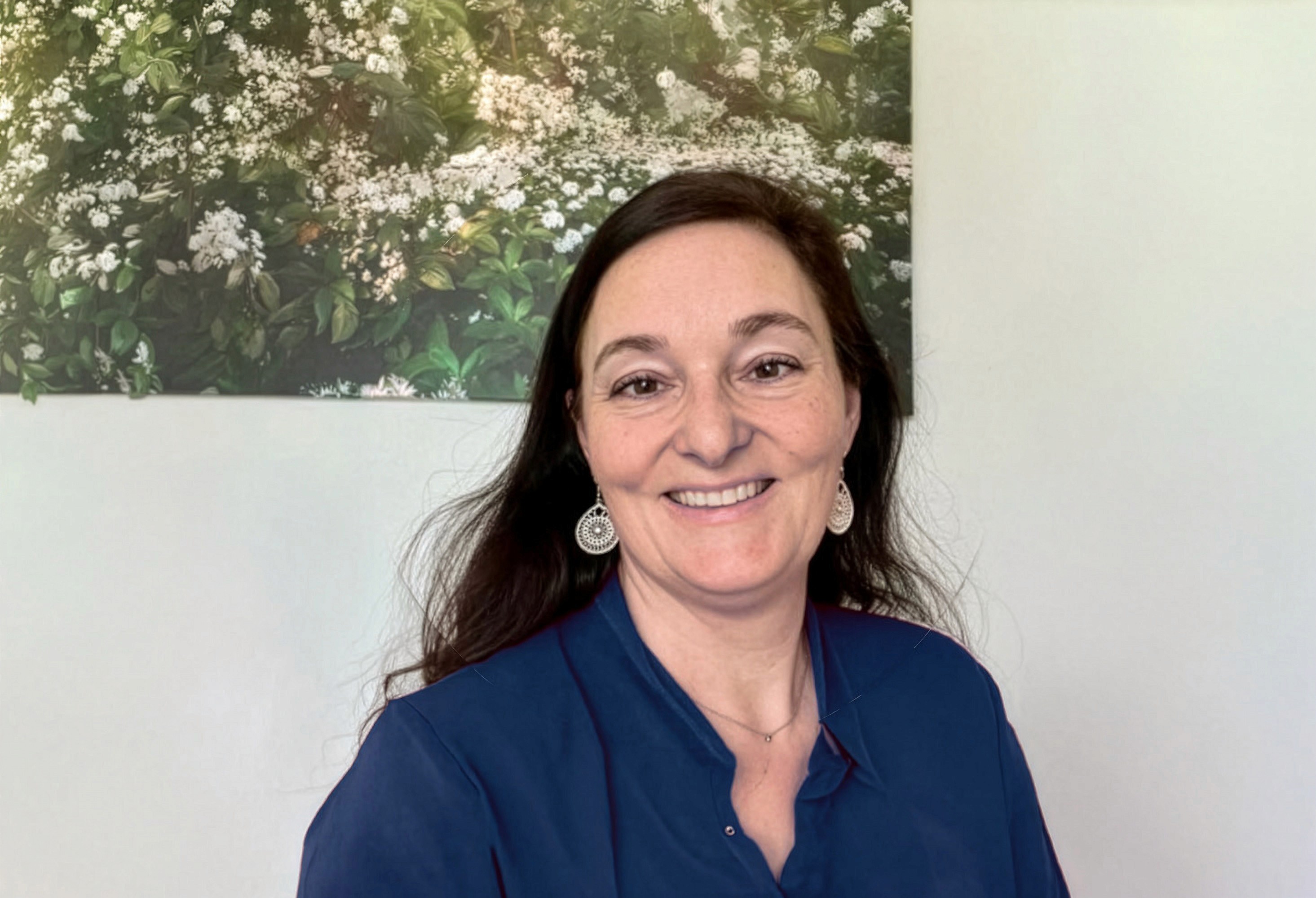 Portrait von Silke Auer, eine Frau mit schwarzen langen Haaren. Sie trägt eine blaue Bluse und steht vo einem Bild.