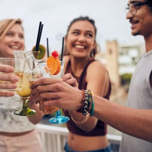 سه دوست با لبخندی بر لب، لیوان‌هایی پر از نوشیدنی‌های رنگارنگ و تزئین شده با میوه را در فضای باز و در یک روز آفتابی به یکدیگر چفت می‌کنند.