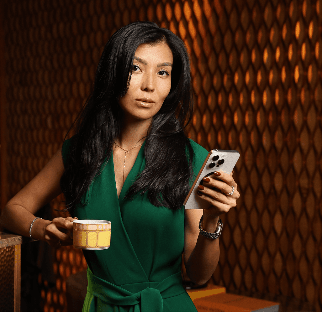 A woman in a green dress holds a cup in one hand and a phone in the other, smiling as she enjoys her drink.