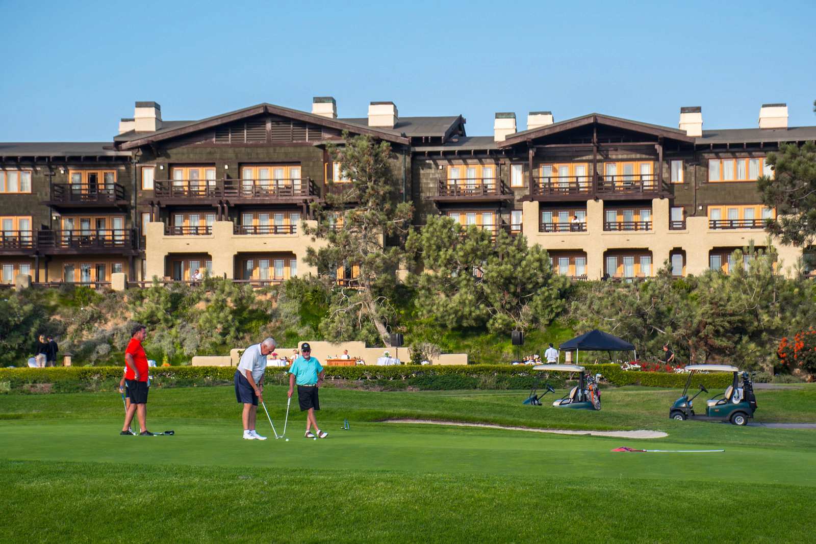 Golfers and a child on a green in front of a multi-story resort building with arched windows under a clear sky.