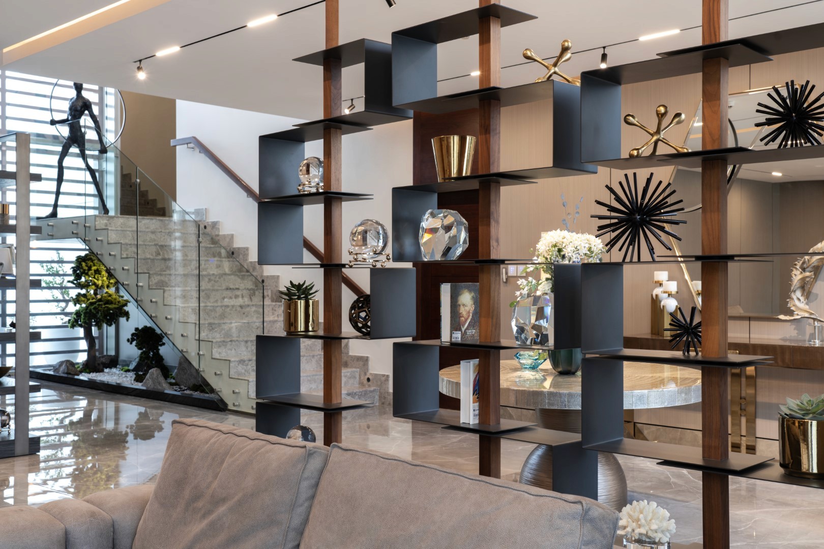 Modern interior of a stylish living room features an open, airy space divided by a geometric black shelving unit filled with artful sculptures and decorative objects, set beside a sleek stone staircase and accentuated by well-placed indoor plants and contemporary lighting.