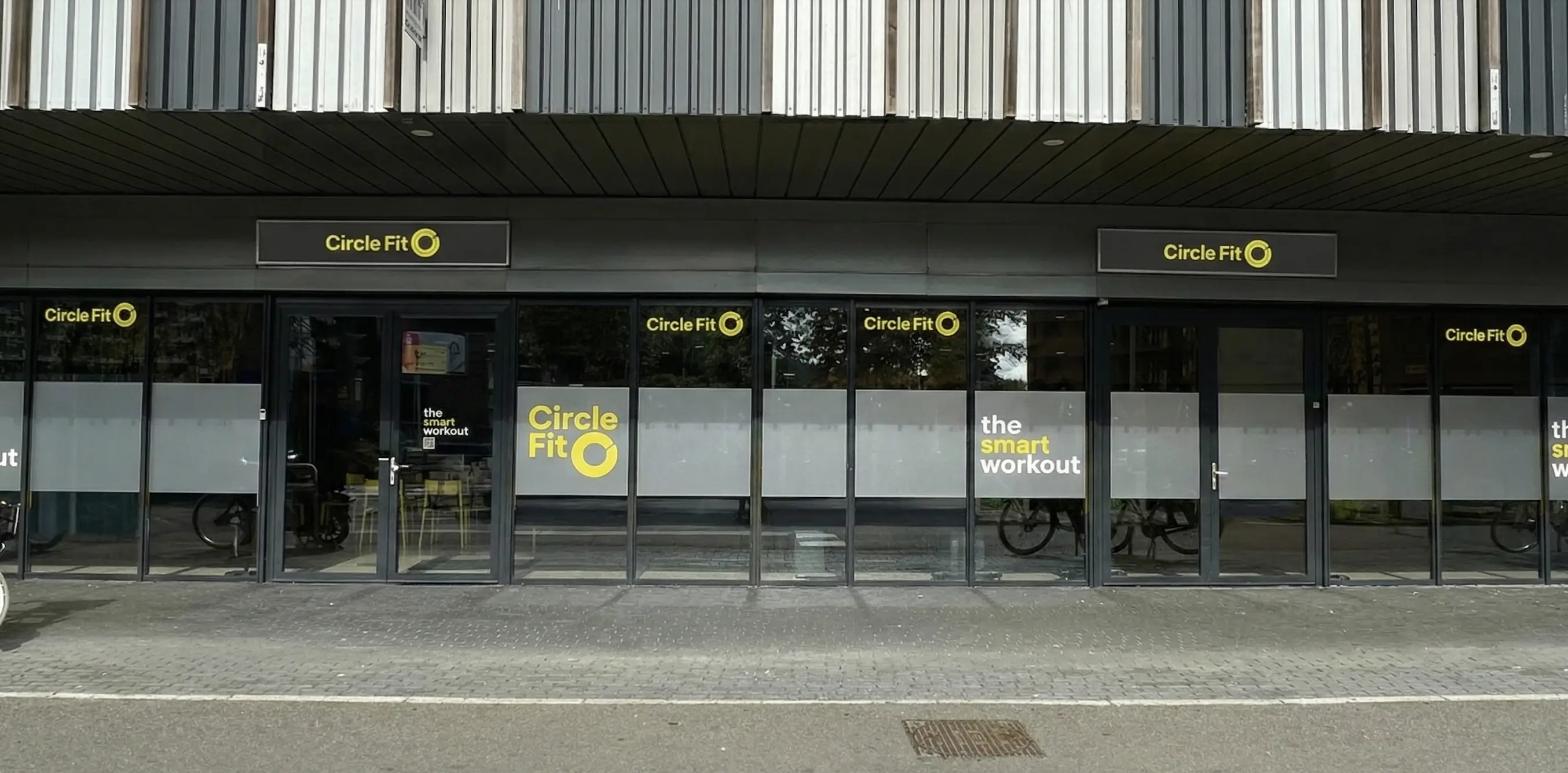 A Circle Fit hub with large glass windows featuring bold yellow signage and people visible indoors. Sidewalk in view.