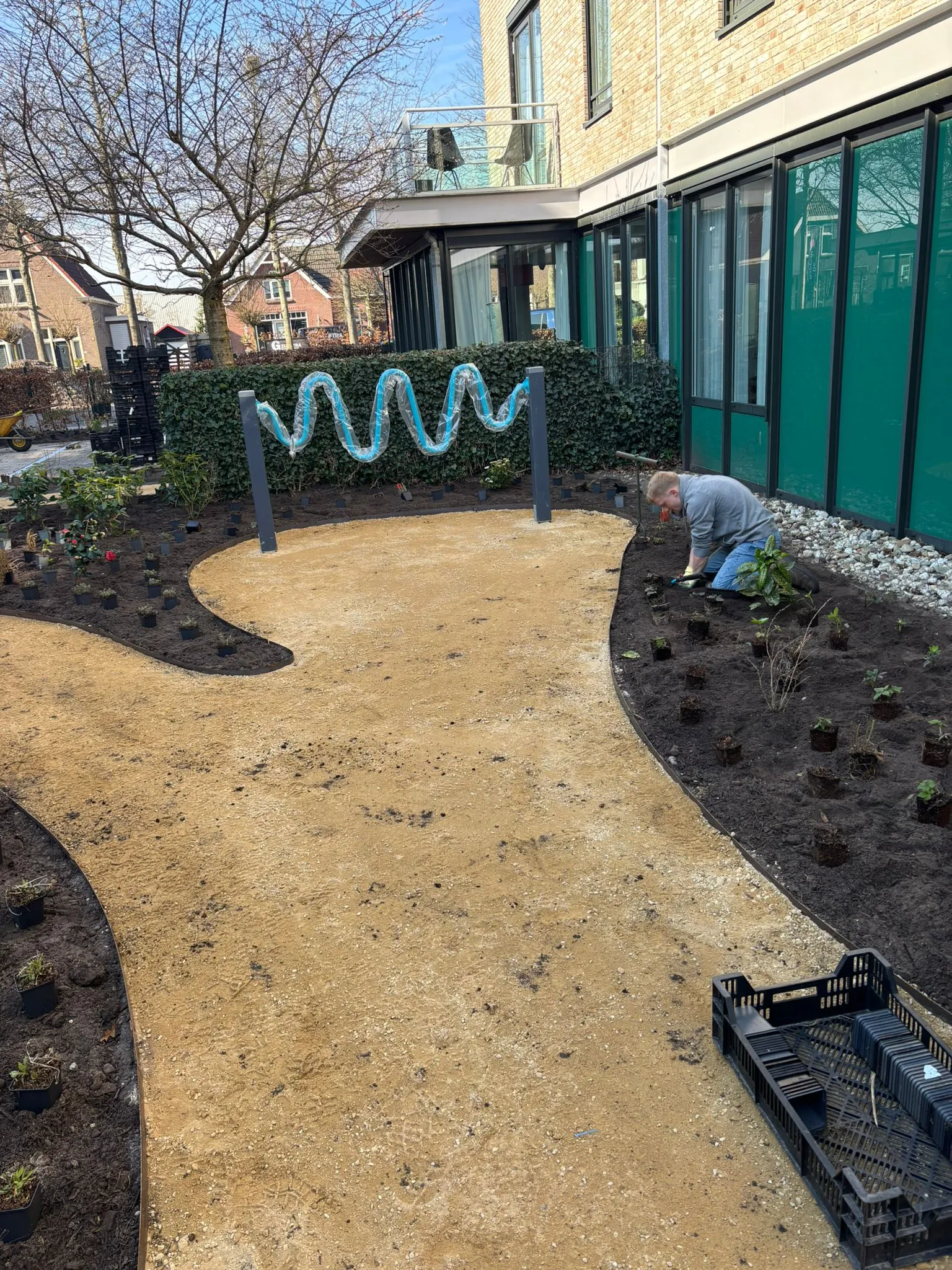 Slingerend zandpad tussen nieuw geplante struiken en planten langs een gebouw, met een persoon die de tuin aanlegt.