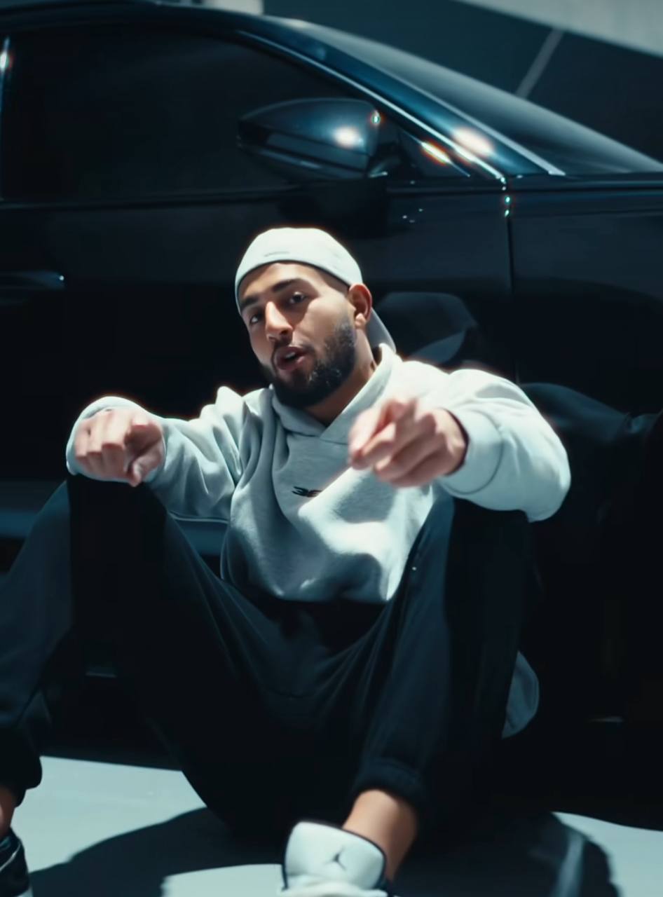 Two people in a workshop, one standing and smiling while the other leans over a car, working on it.
