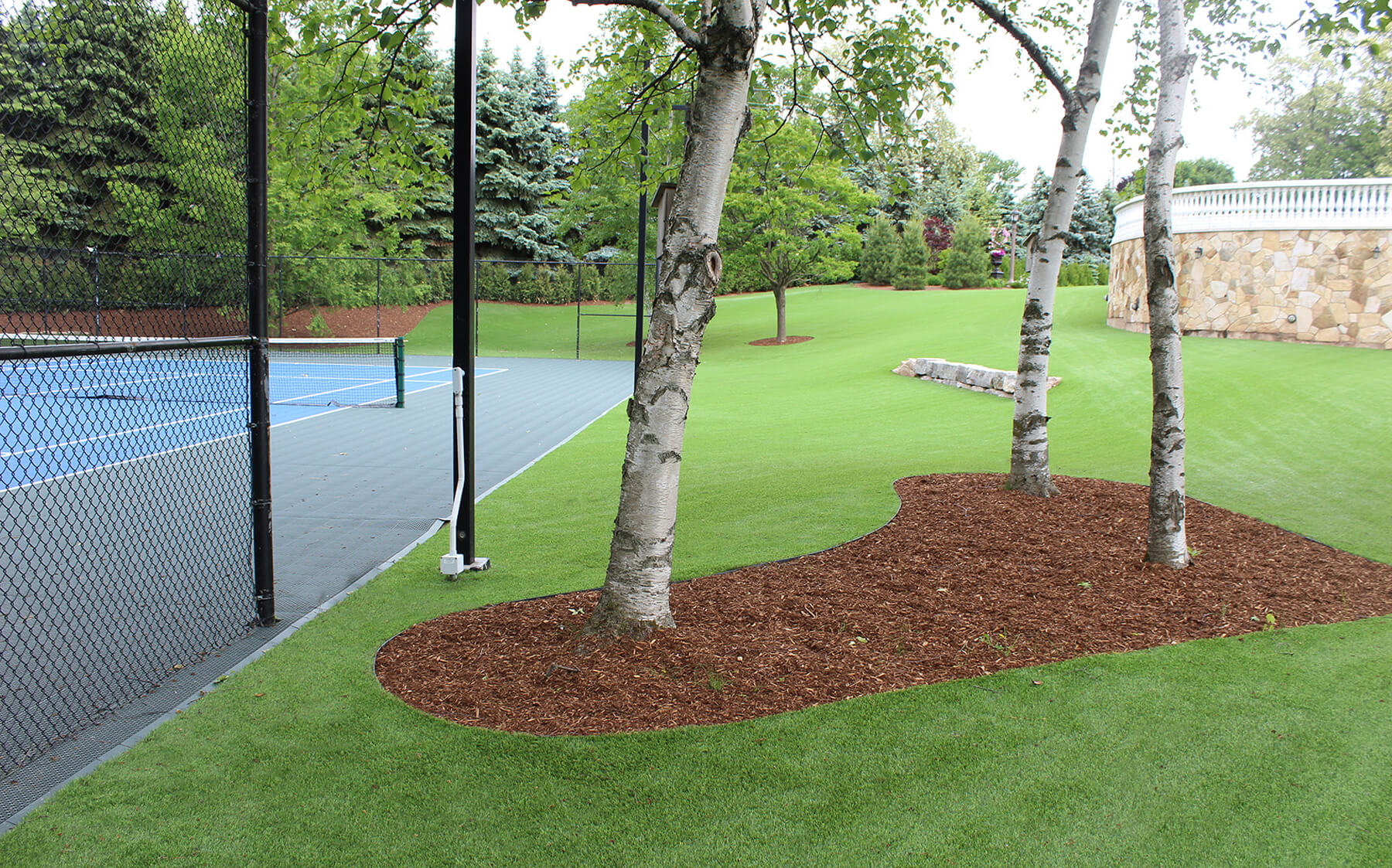 A vibrant tennis court with artificial AGL Grass framed by tall trees, creating a serene outdoor sports environment.
