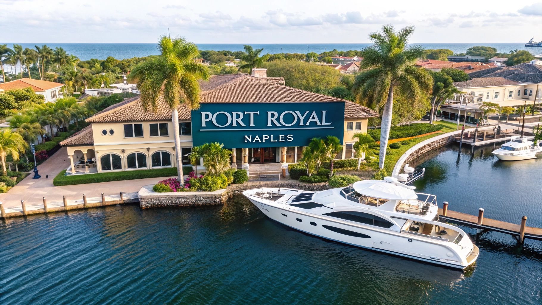  a blue sign reading 'port royal naples' over a large waterfront home with a dock and yacht