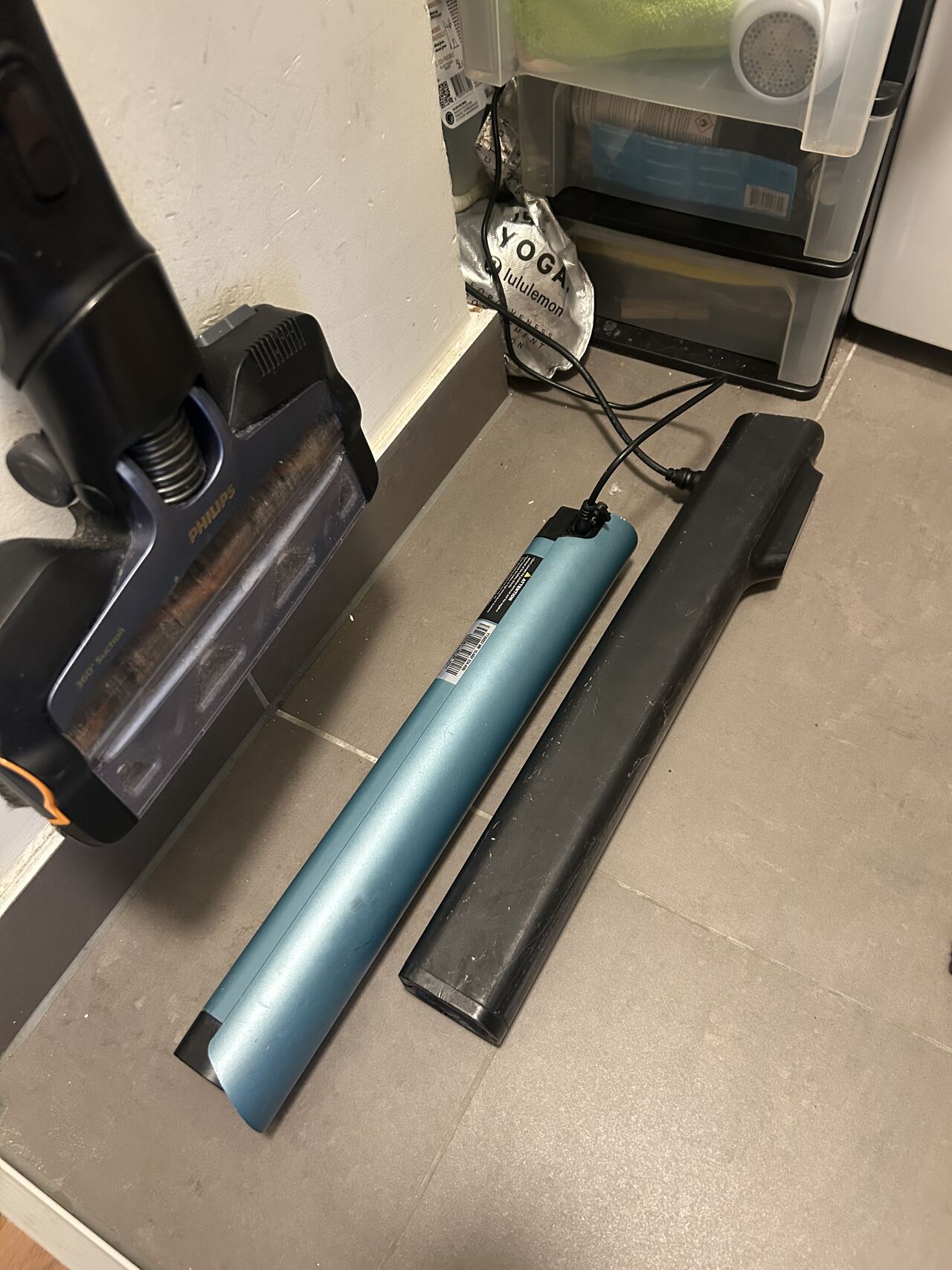 An e-bike battery charging on a utility room floor next to a vacuum cleaner and storage boxes, plugged into a domestic socket.