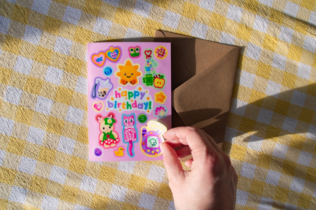 A hand peeling a sticker from a greetings card. The card is pink and has various cute trinket stickers, including a tamagotchi and a sylvanian family rabbit. The centre of the card reads "Happy Birthday!".