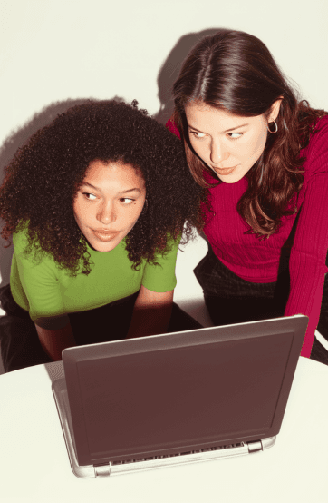 Two women are focused on a laptop screen, one with curly hair wearing green, and the other with straight hair in red.