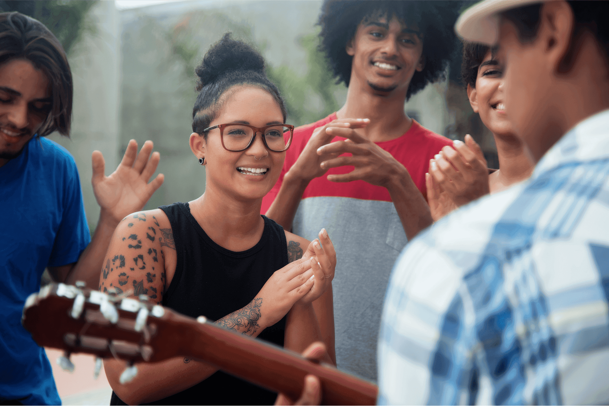A group of young adults is joyfully clapping and smiling outdoors, surrounding a person playing an acoustic guitar, capturing a lively and engaging moment of musical enjoyment and community.