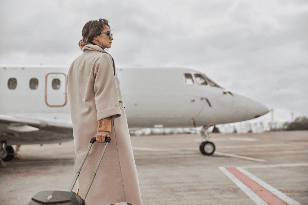 A well-dressed woman in a beige coat and sunglasses pulling a suitcase across an airport tarmac, with a private jet in the background.