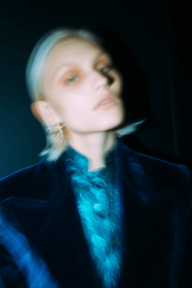 Stylized fashion portrait of a woman wearing a blue outfit and statement earrings, photographed with intentional motion blur against a dark background.
