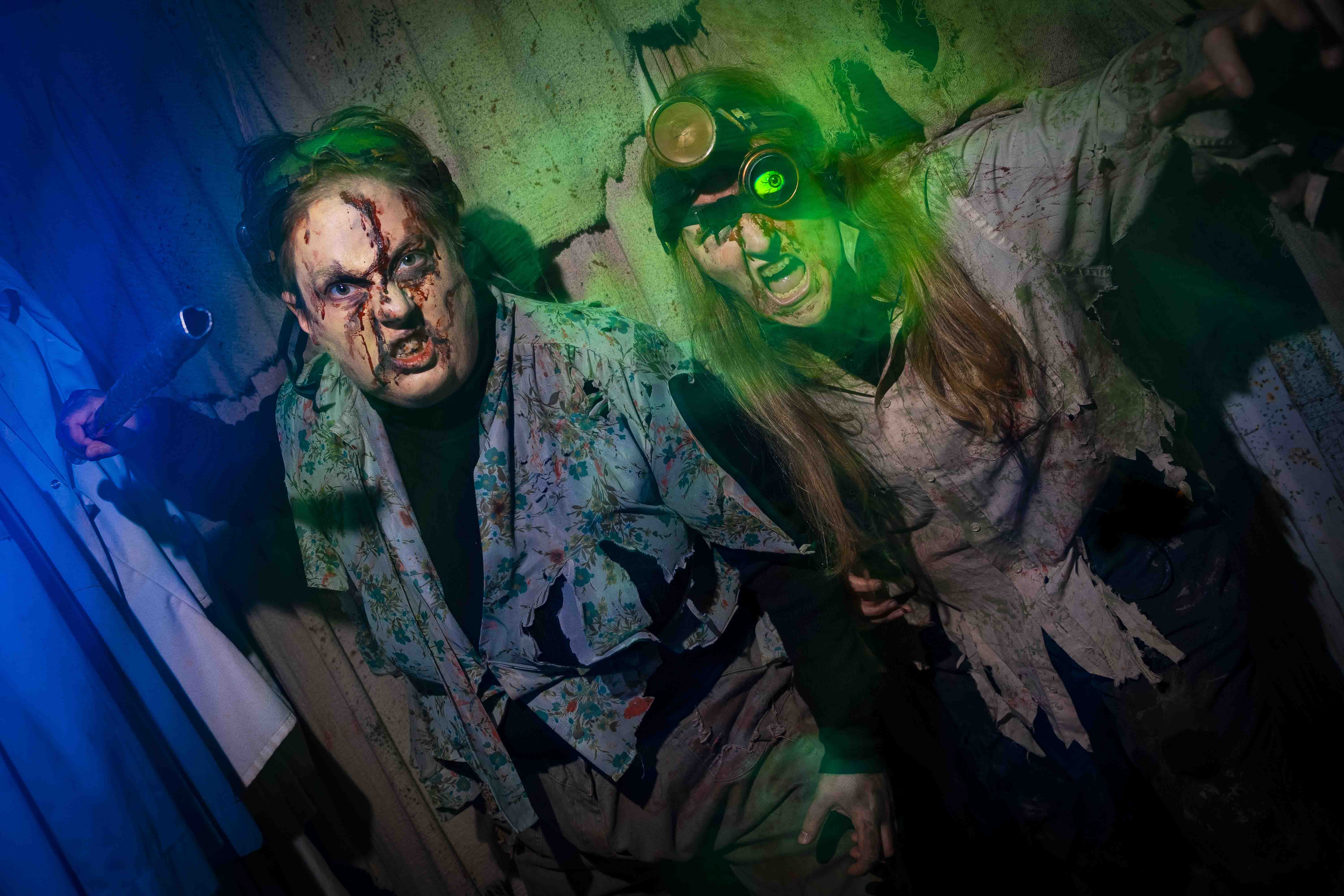 Two actors in zombie costumes, covered in blood and grime, with green lighting effects. The man on the left is holding what looks like a weapon. 