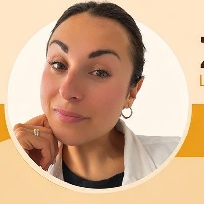 A woman with blonde bob hair in a grey coat and beige turtleneck stands against a neutral backdrop. She has a serious expression, conveying elegance.