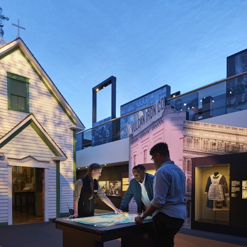 三个人在博物馆展览中查看展示台，周围是历史建筑和文物，环境昏暗。