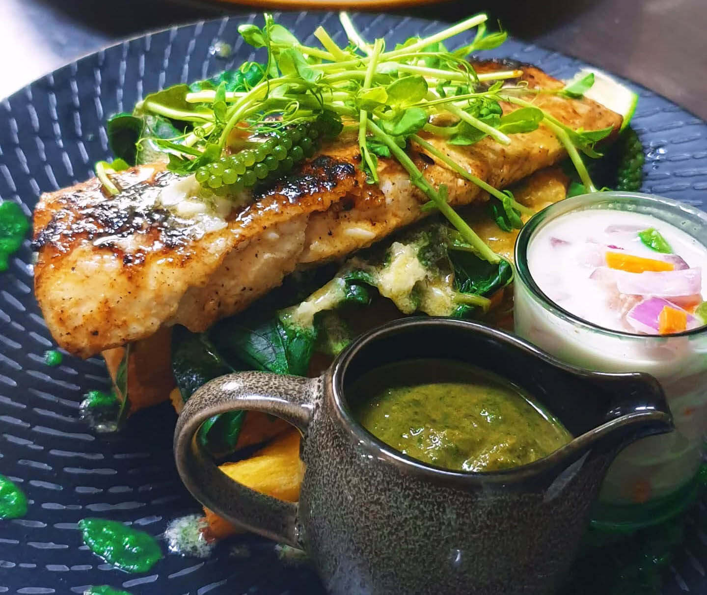 Grilled fish fillet with fresh greens, sprouts, and sea grapes, served with a green sauce and a creamy side dish.