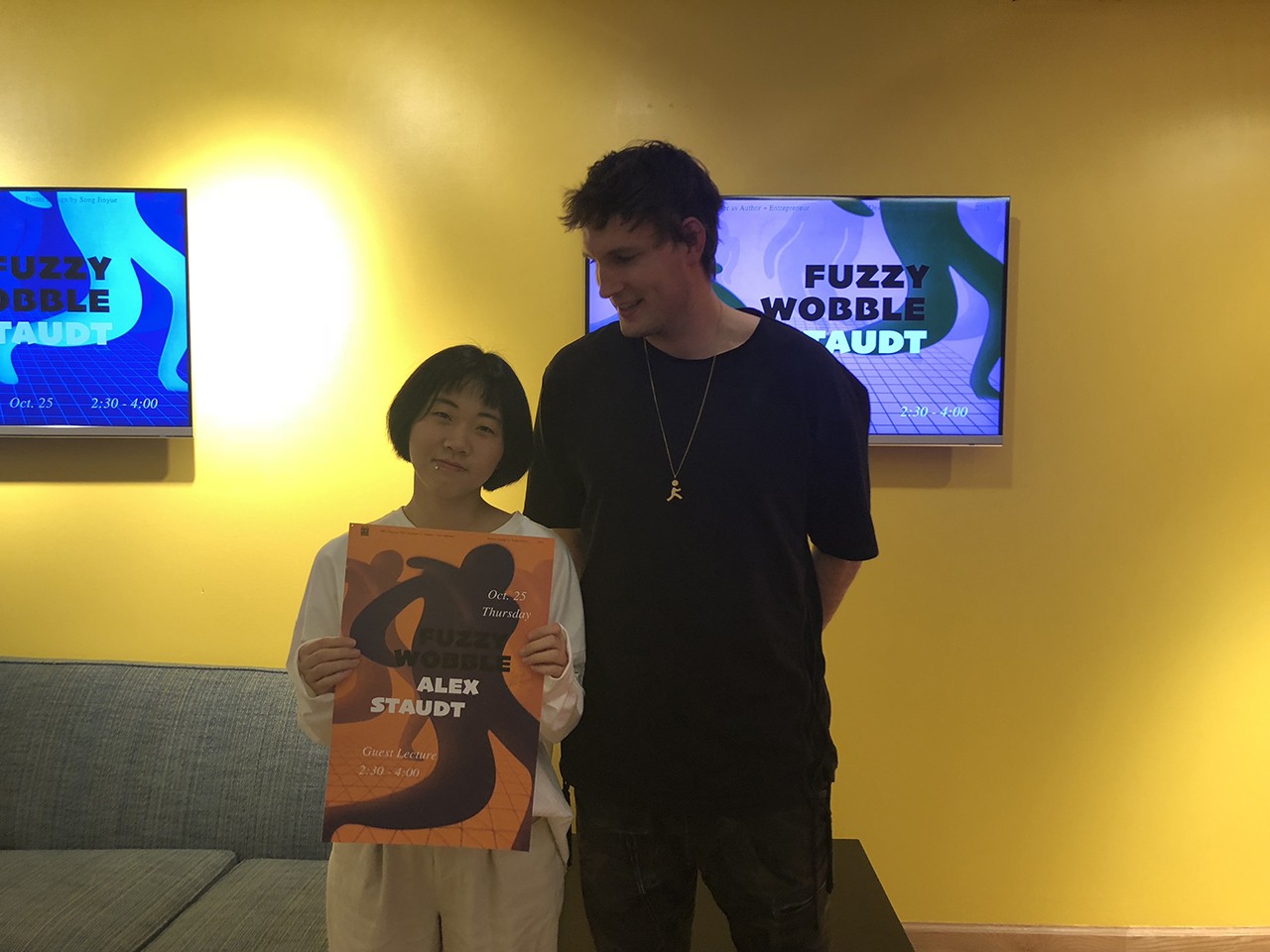 A photo of two people, one holding an orange poster with some figures on it. They both are standing in an exhibition room and behind them on the yellow wall there are some lcd screens with artwork.
