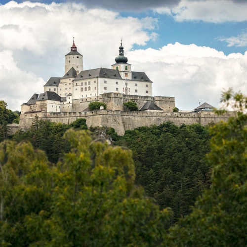 A large castle with multiple towers sits atop a hill, surrounded by dense greenery and under a partly cloudy sky.