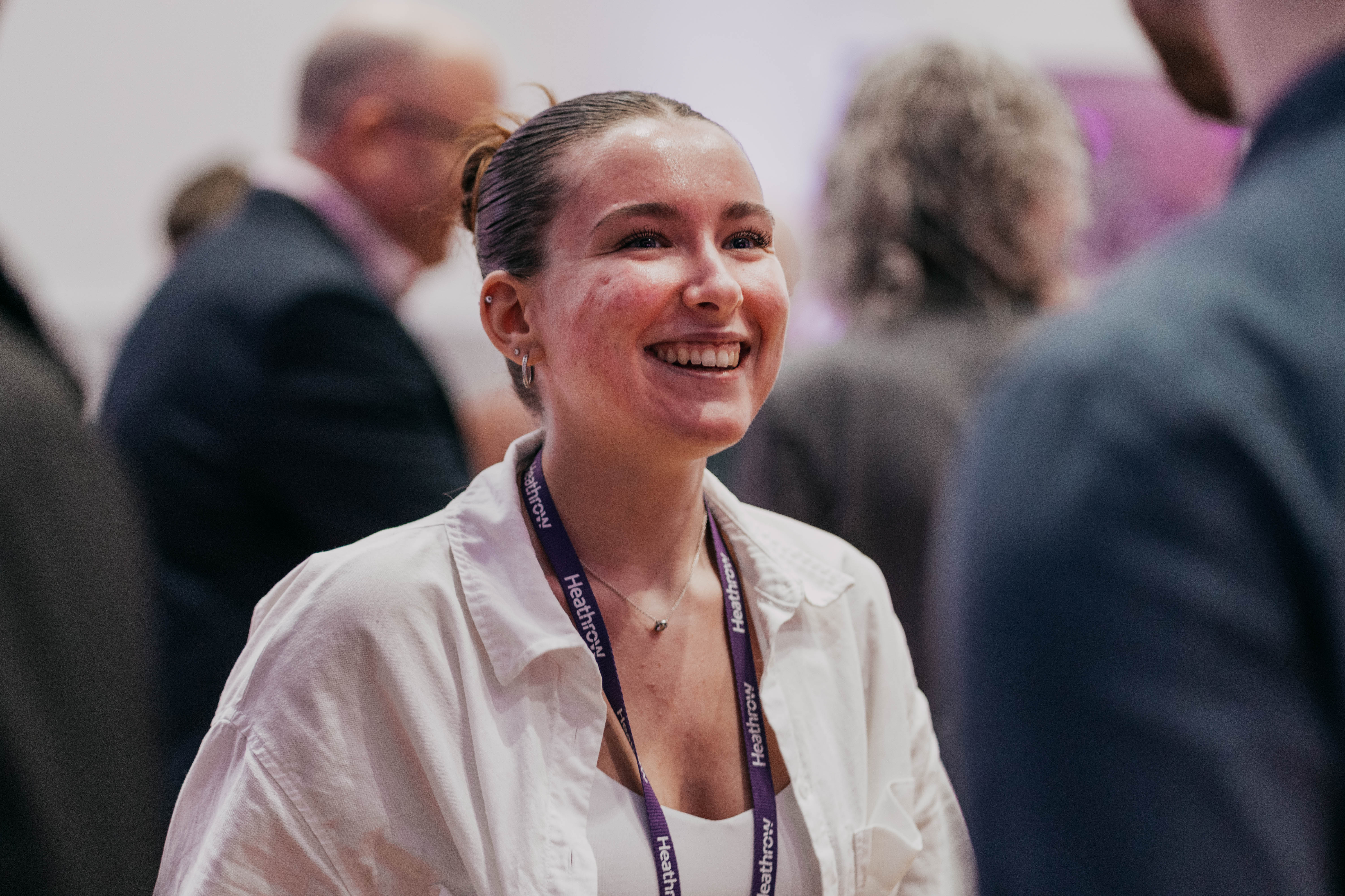 A candid photo of a young woman with her hair pulled back, smiling warmly while wearing a white shirt and a Heathrow lanyard at an event.