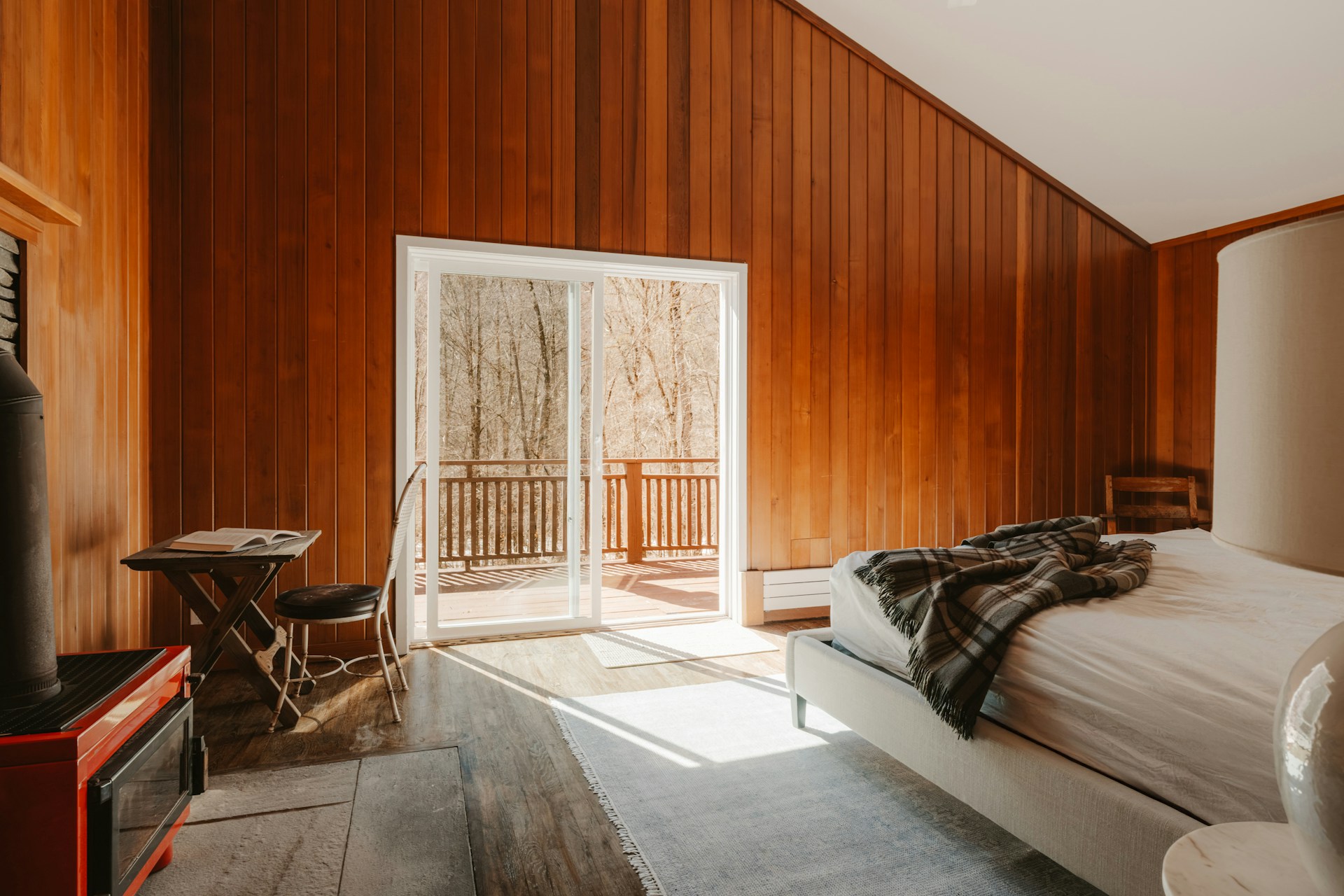 Sunlit, wood-paneled cabin bedroom with a bed, red stove, and glass doors opening onto a wooded deck.