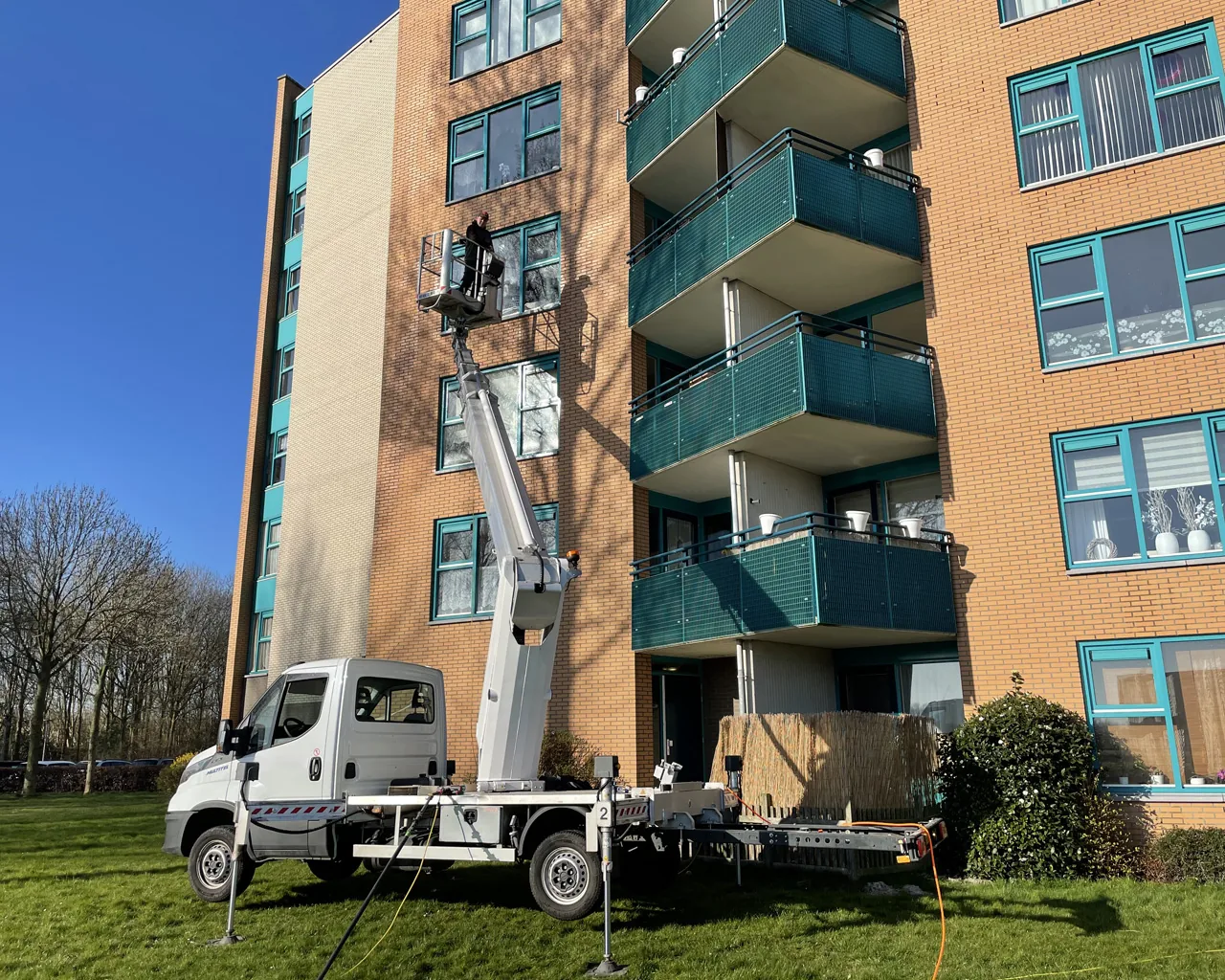 Medewerker in de bak van een vrachtwagenhoogwerker werkt aan de kozijnen van een appartementencomplex met balkons en bakstenen gevel