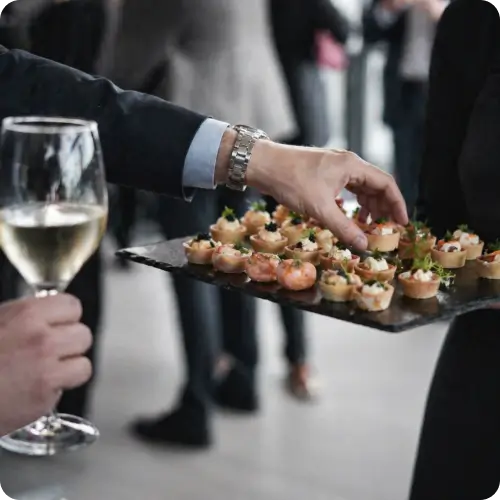 Garçom servindo canapés sofisticados durante coquetel corporativo, com atmosfera elegante e networking ao fundo.