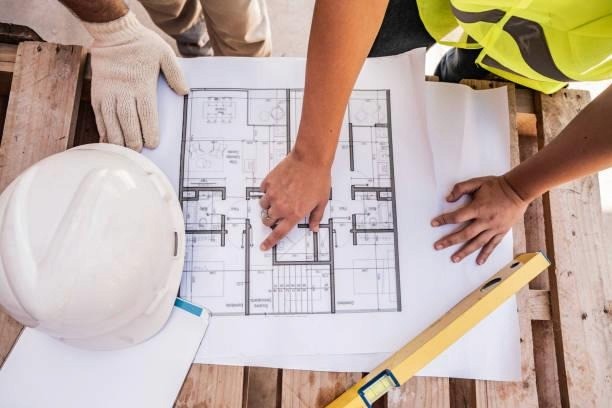 A construction worker wearing a hard hat and high-visibility vest is holding a blueprint and a tape measure, working on a wooden platform.

AI-generated content may be incorrect.