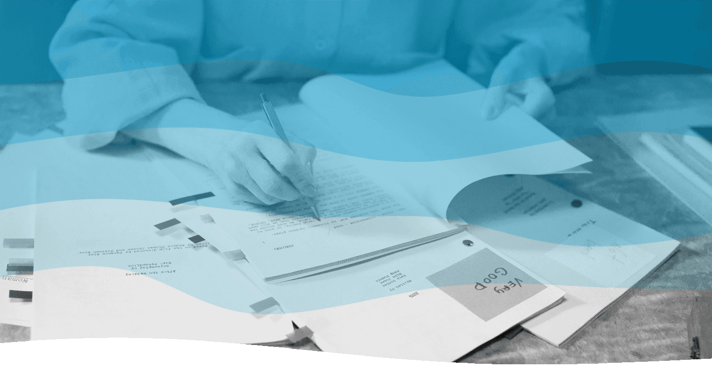 A person is writing with a pen on an open notebook, surrounded by scattered documents and papers on a desk.