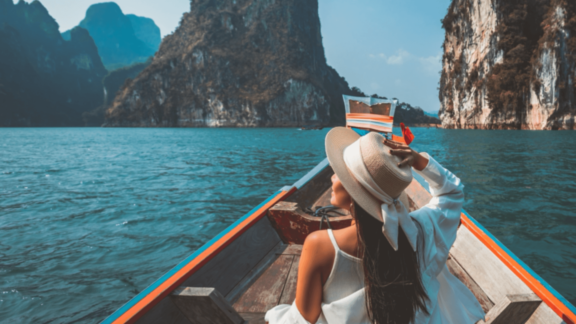 A woman sits in a small, open boat in an exotic location, gazing at the sky and holding onto her hat
