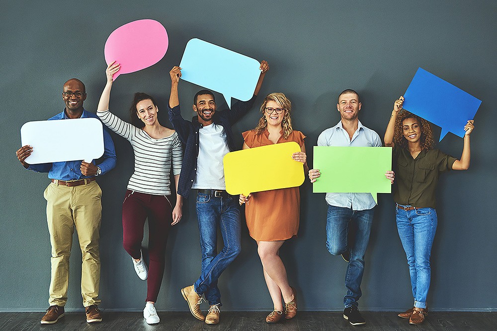Six diverse people standing against a grey wall, each holding a colourful speech bubble cutout and smiling.