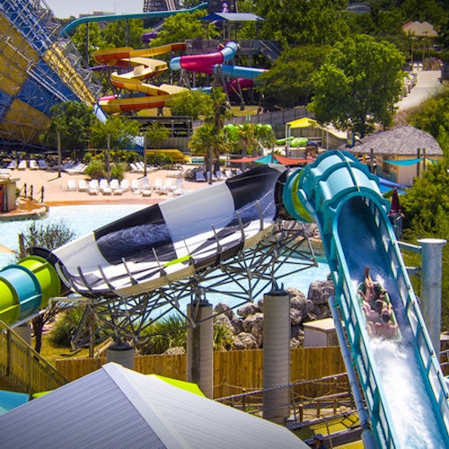 People riding brightly colored water slides at a water park with trees and lounge areas in the background.