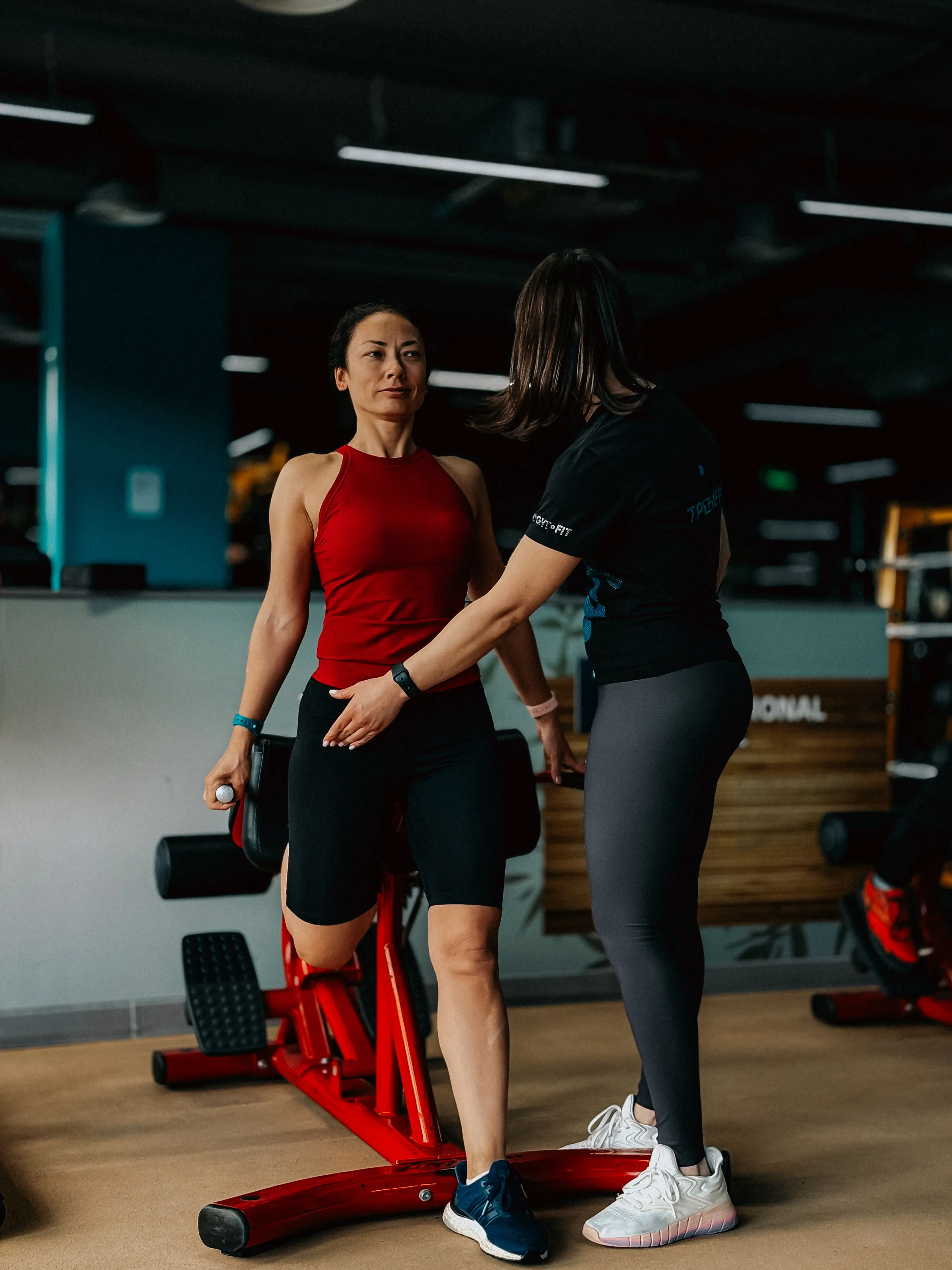 A gym trainer assists a woman in performing a leg exercise on a red exercise machine in a modern gym, both wearing athletic attire and focused on form, surrounded by fitness equipment.