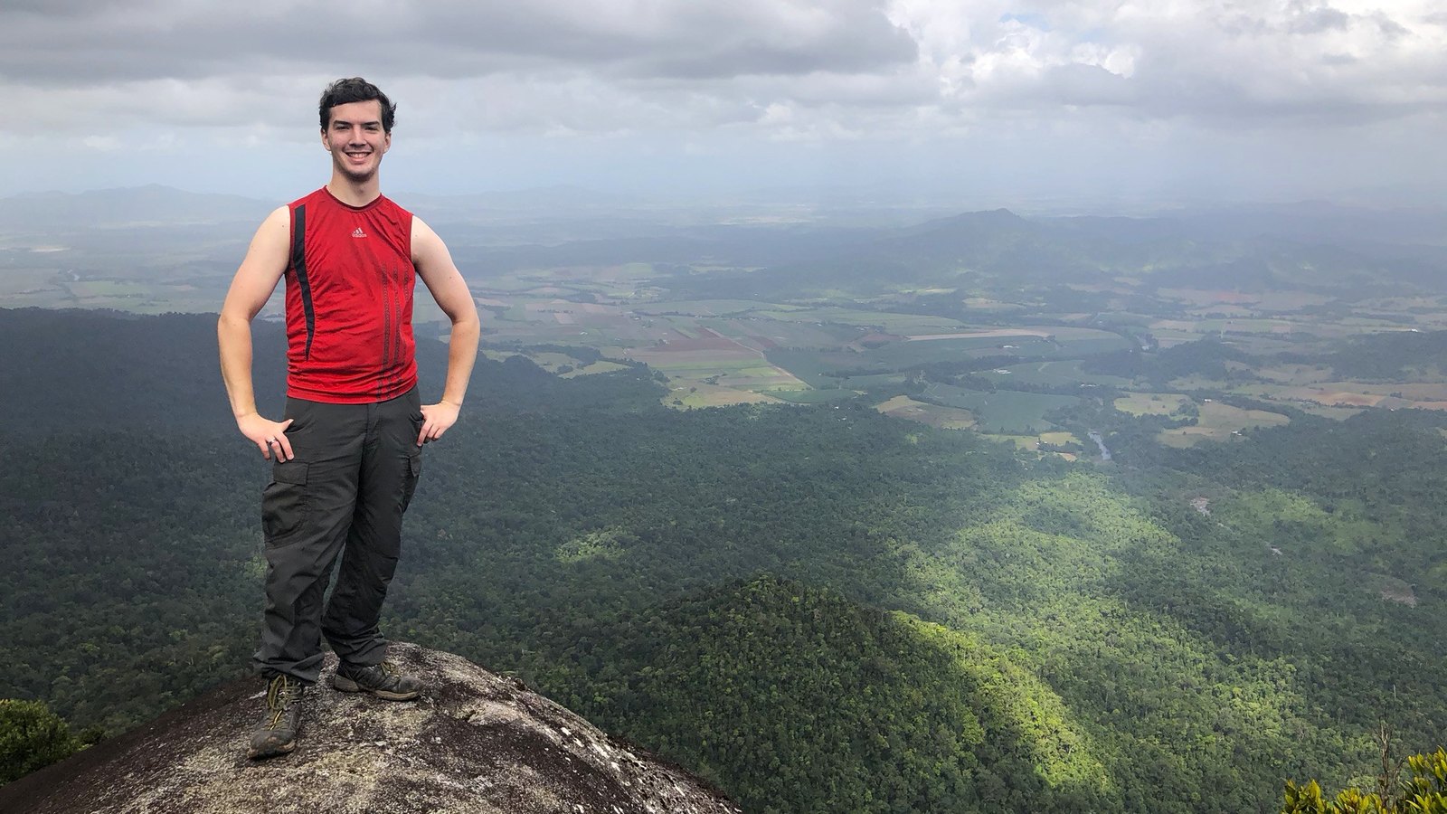 Tim standing on a rocky summit above rainforest and farmland under a cloudy sky.