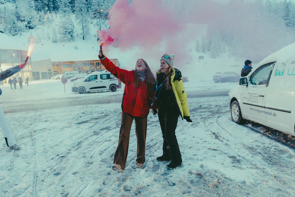 Two women celebrating with pink smoke flares in snowy mountain resort.