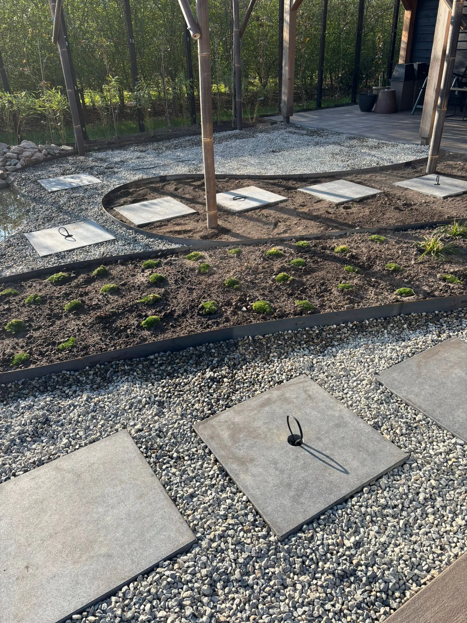 Vierkante betontegels met metalen ringen liggen op grind naast een organisch gevormd tuinbed met jonge planten bij een pergola.