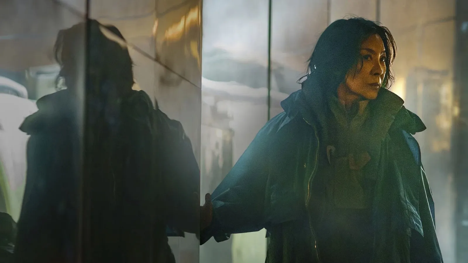 A woman in a dark, reflective room stands with an intense expression, surrounded by mist and wearing a long, heavy coat, which adds to the moody and mysterious atmosphere.