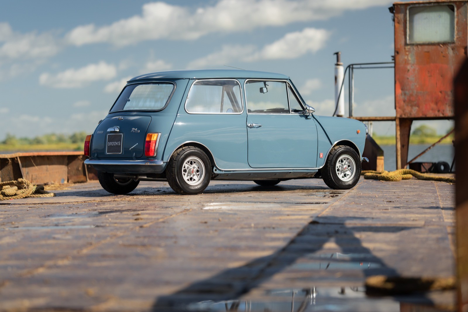 A classic blue car parked on a gravel driveway beside a rustic wooden building under a clear sky.