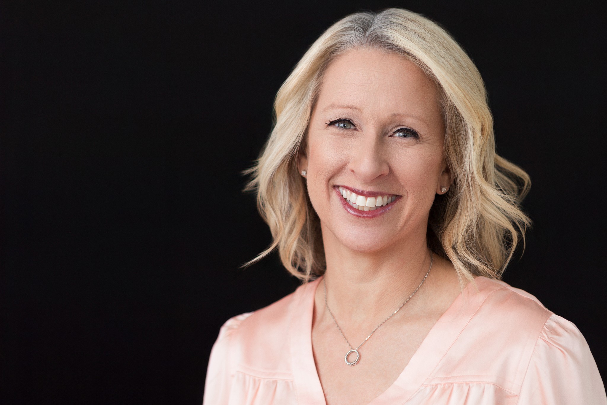 Executive branding portrait of a professional woman smiling against a black background, photographed with soft studio lighting.