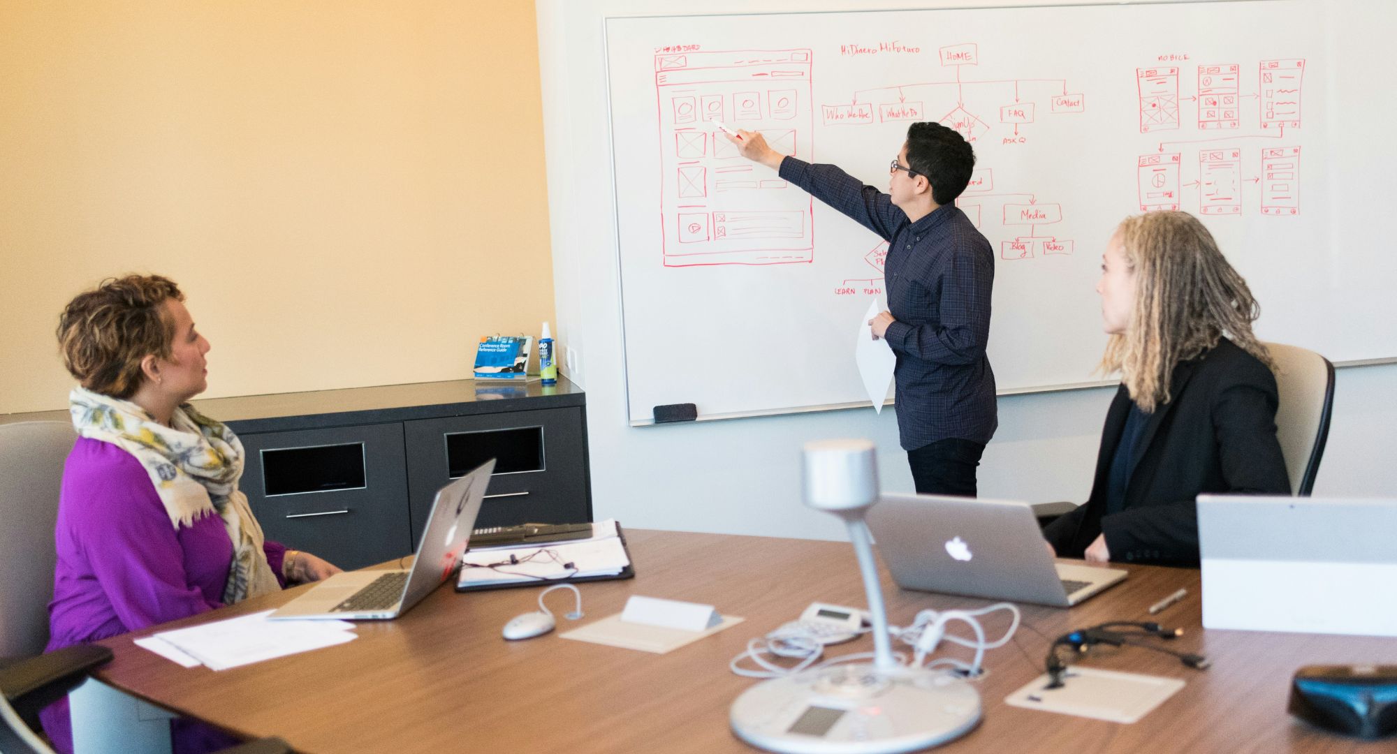 três pessoas numa sala de reuniões, duas mulheres estão sentadas à mesa com notebooks com atenção na terceira mulher de pé escrevendo em um quadro branco
