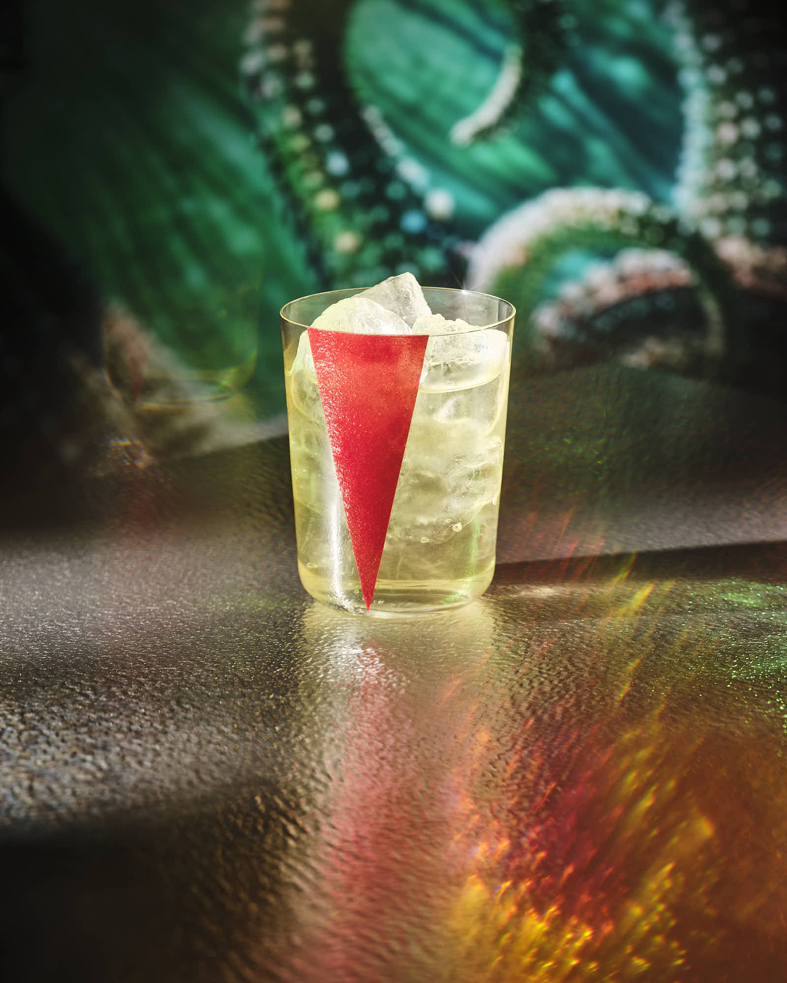 A colorful cocktail glass with a red drink sits on a reflective surface against a vibrant green background.