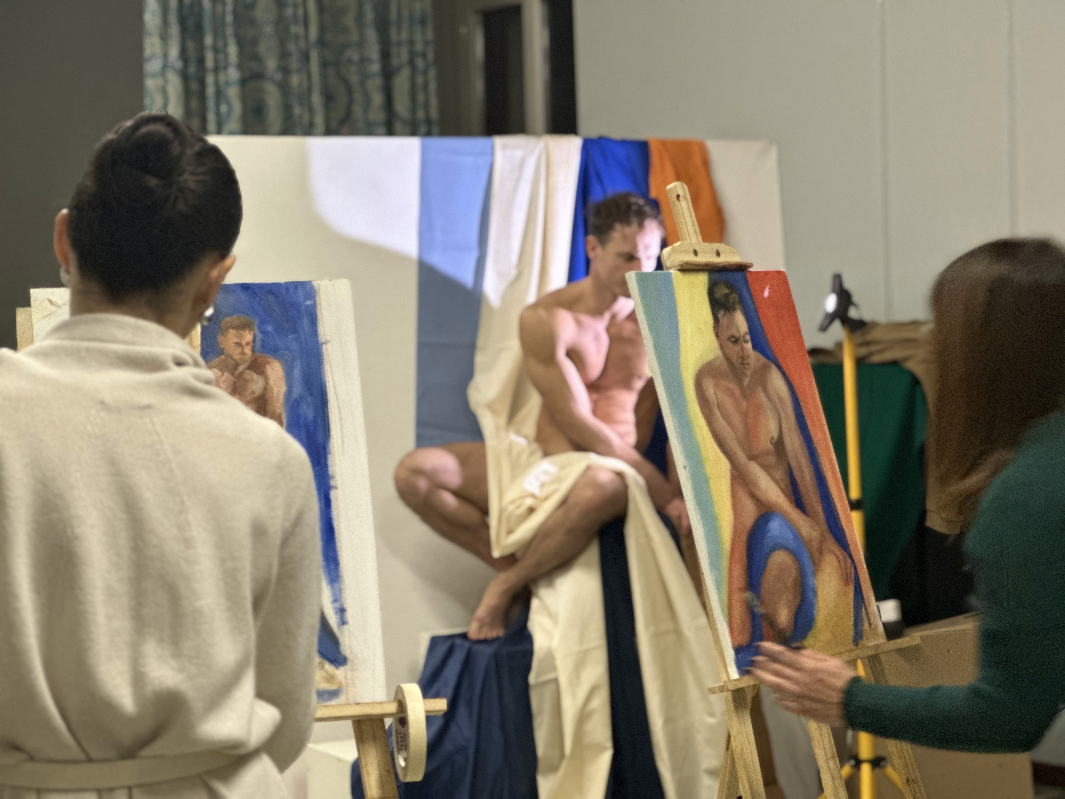 In a well-lit art studio, two artists focus intently on painting a male model posed against a multicolored draped backdrop, emphasizing the creative process and live figure drawing.