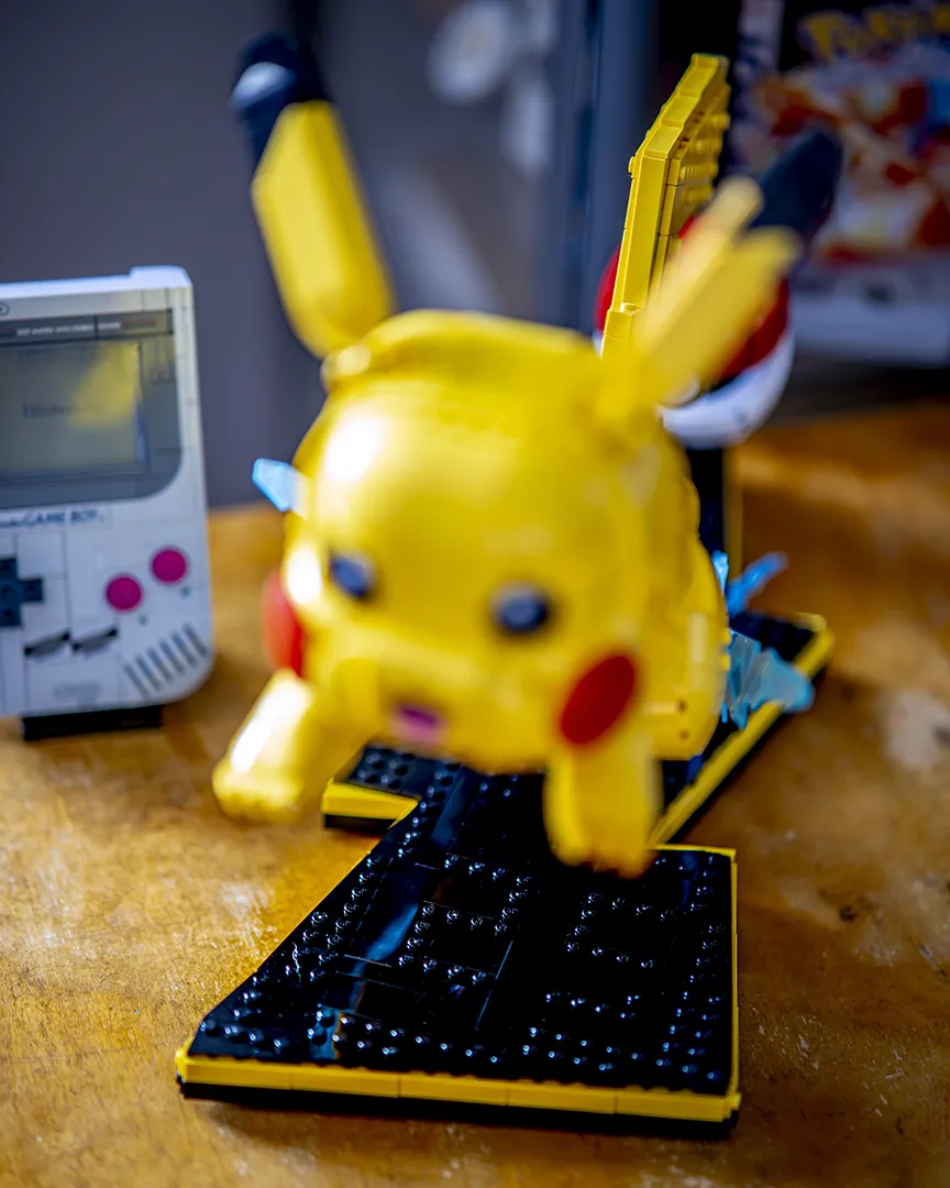 LEGO Pikachu i en dynamisk positur på en svart utstillingsbase, med LEGO Game Boy i bakgrunnen.