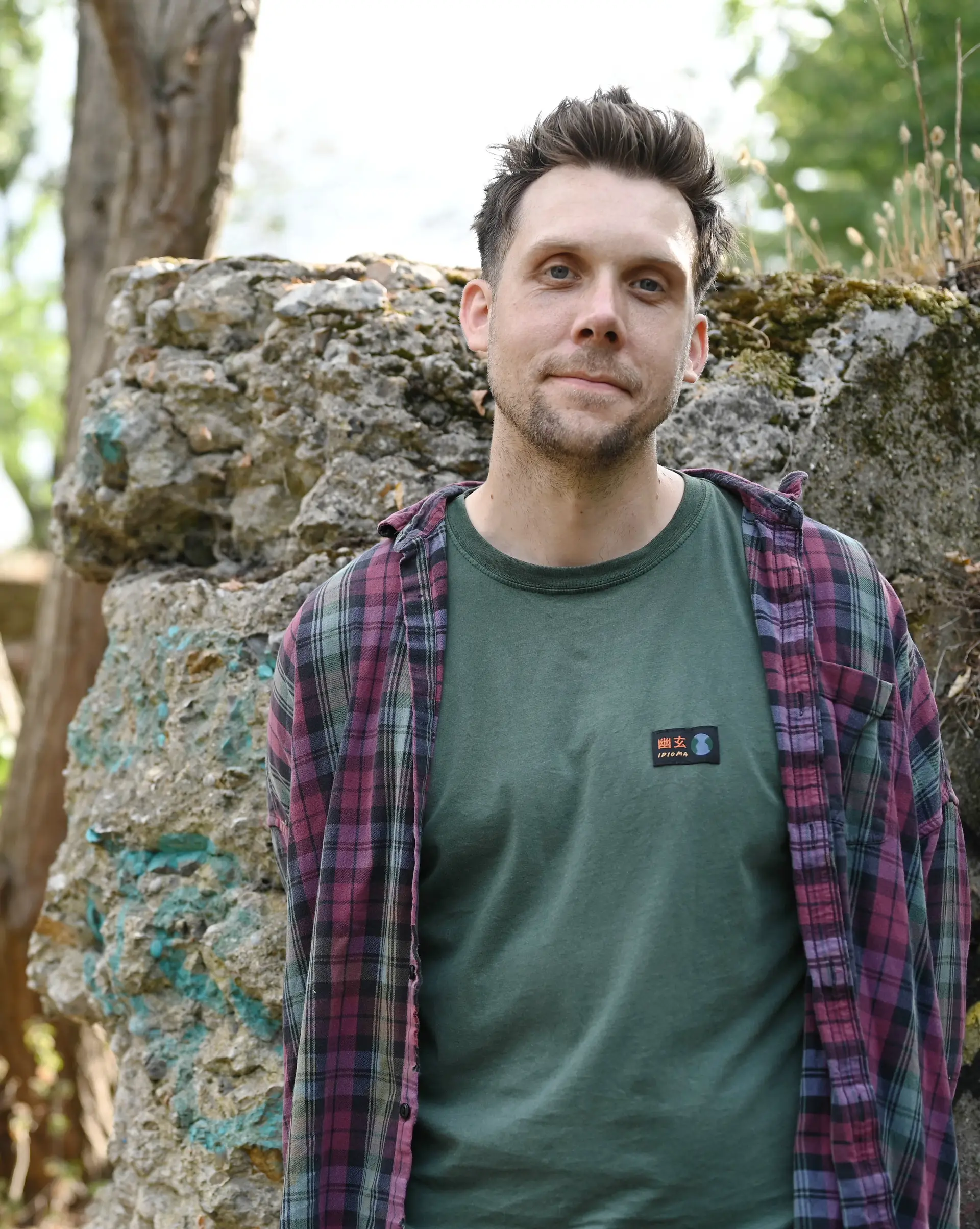 A person wearing a cap and a brown jacket sits outdoors, looking towards the camera with greenery in the background.